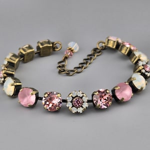 May include: A bracelet featuring round pink and iridescent white crystals set in antique brass metal settings. The design includes a central flower-shaped cluster of small crystals, connected by metal links with an adjustable chain and a small bead.