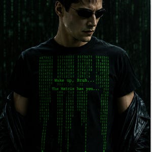 May include: A black short-sleeved t-shirt featuring a green digital code pattern printed on the front. The graphic includes the text "Wake up, Bruh..." and "The Matrix has you..." displayed in a font style reminiscent of computer terminal data.