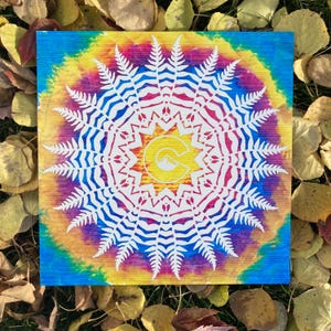 May include: A white fern mandala design on a colorful tie-dye background. The mandala features a stylized letter "C" with a mountain peak inside. The background is a vibrant mix of blue, yellow, red, and green.