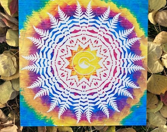 May include: A white fern mandala design on a colorful tie-dye background. The mandala features a stylized letter "C" with a mountain peak inside. The background is a vibrant mix of blue, yellow, red, and green.
