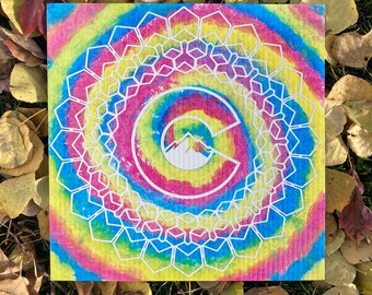 May include: A colorful tie-dye patterned square dishcloth with a white outline of a stylized letter "C" with mountains inside the letter. The dishcloth has a circular pattern of white hexagons.