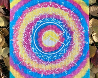May include: A colorful tie-dye fabric with a white outline of a geometric pattern and a white letter "C" with a mountain silhouette inside. The letter "C" is in the center of the fabric.