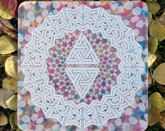May include: A white geometric design on a clear acrylic coaster with a colorful background. The design features a central diamond shape with a repeating pattern of triangles and lines. The background is a mix of pink, blue, and yellow.