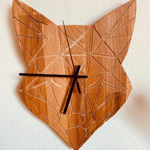 Könnte beinhalten: Eine Wanduhr aus Holz in Form eines Fuchs Kopfes mit geometrischem Design. Die Uhr hat schwarze Zeiger und ein weißes Zifferblatt.