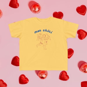 May include: A yellow t-shirt featuring a graphic of a cupid with the text "mon chéri" in blue and red.