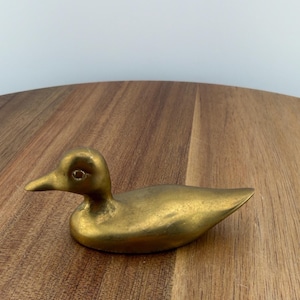 May include: A small, brass duck figurine. The duck is in a resting position, with its head turned slightly to the side. The brass has a warm, golden hue. The duck is a decorative item.