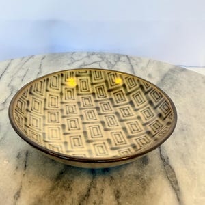 May include: A shallow ceramic bowl featuring a repeating geometric pattern of dark brown squares and rectangles on a light beige background. The bowl has a smooth, rounded rim with a dark brown edge that contrasts with the interior design.