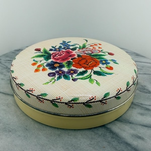May include: A round, vintage tin with a floral design on the lid. The lid features a cream-colored background with a colorful bouquet of red, pink, blue, and purple flowers. The tin has a yellow base and a silver rim.