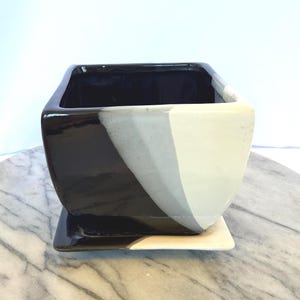 May include: A square ceramic planter with a glossy finish, featuring a color-blocked design in black, grey, and white. The planter includes a matching square saucer base, with the black and white sections extending onto the tray surface.