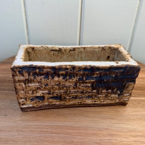 May include: A rectangular ceramic planter with a textured brick pattern. The planter has a brown and white color scheme, with darker brown accents around the rim. The interior is a lighter beige color. The planter sits on a wooden surface.