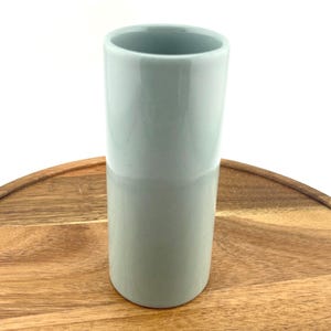 May include: A two-tone ceramic tumbler in shades of light blue and gray. The cylindrical cup is set on a wooden tray. The top half of the tumbler is light blue, while the bottom half is gray. The tumbler is approximately 6 inches tall.