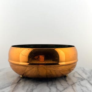 May include: A copper-colored bowl with a rounded shape and a dark interior. The bowl sits on three small, gold-colored feet. The surface of the bowl reflects the surrounding environment. The bowl is likely used for decorative purposes.