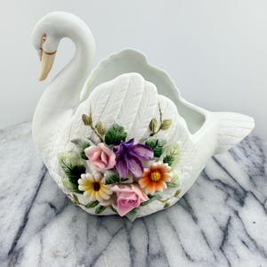 May include: A white ceramic swan-shaped planter with a detailed design. The swan features a curved neck and a beak with a touch of gold. The planter is adorned with colorful, three-dimensional flowers, including pink roses, and purple and orange blooms.