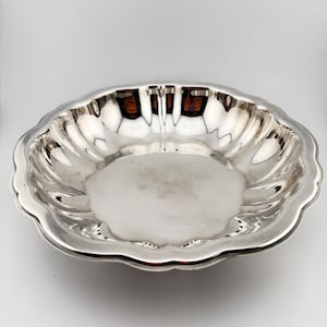 May include: A silver-colored, scalloped-edge bowl. The bowl has a wide, shallow design with a smooth, reflective interior. The rim of the bowl has a wavy, decorative edge. The bowl is likely made of metal and is set against a white background.