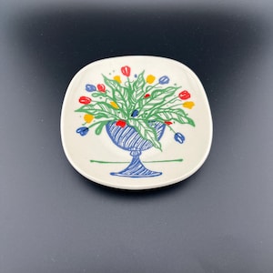 May include: A small, off-white ceramic plate with a hand-drawn floral design. The illustration features a blue vase filled with green leaves and red, yellow, and blue flowers. The plate has rounded corners and a square shape.