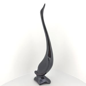May include: A tall, slender, black sculpture featuring a smooth, glossy finish. The object has an elongated, curved neck that tapers to a point at the top, resting on a wide, stable base. The form is abstract, resembling a stylized bird or swan.