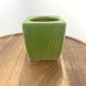 May include: A small, square, green ceramic planter with vertical textured lines on the sides. The planter has a square opening at the top and is set on a wooden surface. Ideal for small plants or succulents.