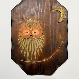 May include: A string art wall hanging on a dark stained wooden plaque with scalloped edges. The design features an owl created with orange and yellow thread, perched on a thin branch, with a yellow crescent moon in the upper right corner.