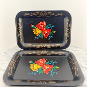 May include: Two black rectangular trays with floral designs. Each tray features a bouquet of red and yellow roses with blue accents. Gold scrollwork borders the edges of the trays, adding a decorative touch to the serving pieces.