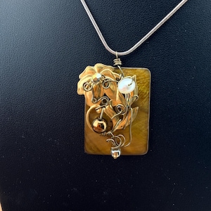 May include: A rectangular tiger's eye pendant on a thin silver-tone chain. The pendant features a gold-tone metal floral accent, intricate wire wrapping, a single white pearl, and a small metallic sphere, all secured to the stone surface.