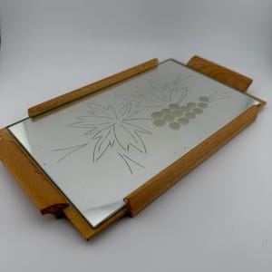 May include: Rectangular mirrored tray with etched floral and grape design. The tray has a wooden frame and handles. The mirror surface reflects light, and the wooden frame has a natural finish. A decorative serving piece.