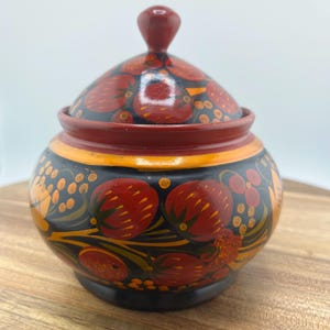 May include: A hand-painted, round wooden trinket box with a lid. The box is black with red and orange floral designs. The lid and base are a dark red color. The box is sitting on a wooden surface.