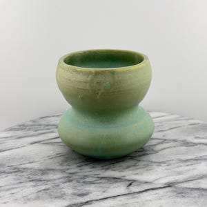 May include: A handmade ceramic vase with a unique hourglass shape. The vase features a two-tone color scheme, with a light green top and a turquoise-green base. The surface has a textured, matte finish, adding to its artisanal appeal. The vase is a decorative item.