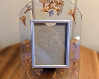 May include: A clear, arched photo frame with a silver border. The frame is decorated with dried flowers in shades of brown and purple. The frame is sitting on a wooden surface.