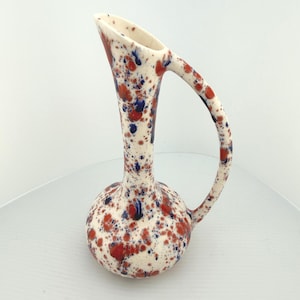 May include: A ceramic pitcher with a tall, slender neck and a rounded base, featuring a cream-colored glaze speckled with vibrant red and dark blue paint splatters. The vessel includes a single curved handle extending from the rim to the body.