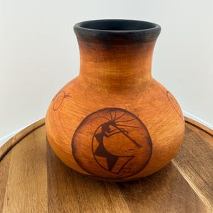 May include: A round terracotta-colored clay vase with a wide body and a narrow neck. The rim is painted black. The exterior features a dark brown circular graphic depicting a stylized flute player, along with smaller circular line drawings.