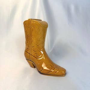 May include: A glossy, mustard-yellow ceramic cowboy boot figurine featuring embossed flame-like decorative stitching patterns on the shaft and toe. The boot has a pointed toe, a stacked heel, and a smooth, reflective finish throughout the surface.