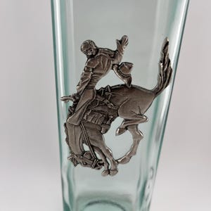 May include: A clear glass bottle with a silver-colored metal emblem of a cowboy riding a bucking bronco. The cowboy is wearing a hat and is holding onto the horse. The bottle has a rectangular shape.