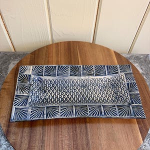 May include: Rectangular ceramic serving dish with a fish scale pattern in the center and a geometric fan design border. The dish is primarily blue and white, with a textured surface. The dish is placed on a wooden surface.