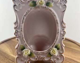 May include: A pink ceramic picture frame with an oval opening. The frame has a decorative design with gold trim and small white rose accents. The frame is sitting on a wooden surface.