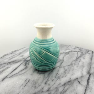 May include: A small, decorative vase with a turquoise body and a cream-colored rim. The turquoise section features a textured, crisscross pattern. The vase is likely ceramic and sits on a marble surface.