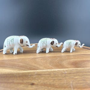 May include: Three white ceramic elephant figurines of varying sizes. Each elephant has brown ears, a trunk, and decorative green and pink patterns on its back. The elephants are displayed on a wooden surface against a dark background.