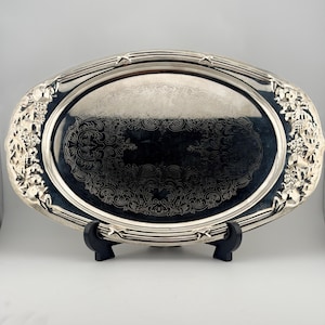 May include: Oval silver serving tray with ornate floral and scrollwork detailing. The tray has a reflective surface and is supported by a black stand. The edges are embellished with relief floral designs.