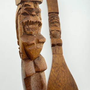 May include: Two wooden kitchen utensils with carved Tiki-style figures. One is a spatula with three slots, and the other is a serving spoon. Both are made of a dark brown wood and have a detailed, carved design.