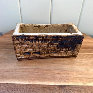 May include: A rectangular ceramic planter with a textured brick pattern. The planter has a light brown exterior with darker brown accents and a white interior. The planter is sitting on a wooden surface.