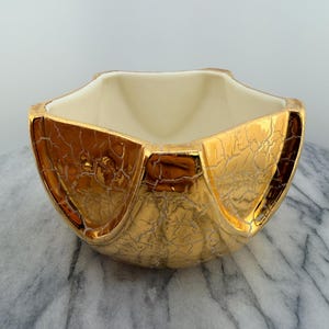 May include: A bowl with a metallic gold exterior featuring a distinct crackle finish and a smooth, off-white interior. The vessel has a wide, open top with a geometric, multi-sided rim and a rounded base, showcasing a reflective, textured surface.