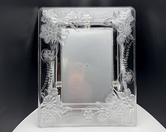 May include: A rectangular clear glass picture frame featuring a frosted, etched floral design. The border is decorated with embossed flowers, leaves, and delicate swirling vine patterns that extend along the sides of the frame, surrounding a central rectangular opening.