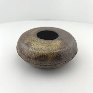 May include: A small, round, ceramic vase with a dark brown exterior and a circular opening. The vase has a textured surface and a dark interior. The color is a mix of brown and gold.