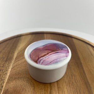 May include: A small, round, off-white ceramic dish with a lid. The lid features a colorful abstract design in shades of pink, purple, and brown. The dish is sitting on a wooden surface.