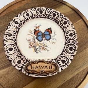 May include: A decorative plate featuring a butterfly with blue and orange wings, set against a floral background. The plate has a brown and cream-colored ornate border and the word "HAWAII" is displayed at the bottom.