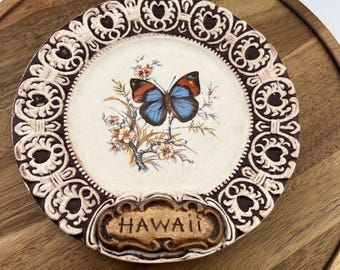 Könnte beinhalten: Ein dekorativer Teller mit einem Schmetterling mit blau-orangen Flügeln vor einem floralen Hintergrund. Der Teller hat einen braunen und cremefarbenen verzierten Rand und das Wort "HAWAII" ist unten abgebildet.