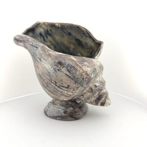 May include: A ceramic vessel shaped like a conch shell, featuring a mottled glaze with shades of grey, brown, and cream. The surface has a textured, weathered appearance with dark streaks and speckles throughout the exterior and interior.