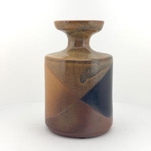 May include: A ceramic vase with a geometric design. The vase features a cylindrical body with a flared top and a dark brown, tan, and black color scheme. The design is composed of angular shapes.