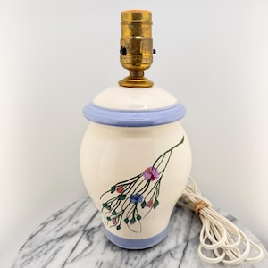 May include: A cream-colored ceramic lamp with a floral design. The lamp has a light blue rim and a brass lamp holder. A white cord is attached to the lamp. The lamp is sitting on a marble surface.