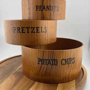 May include: A tiered wooden serving tray with three bowls labeled "PEANUTS", "PRETZELS", and "POTATO CHIPS". The bowls are stacked on top of each other, with the largest bowl at the bottom. The tray is made of wood.