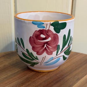 May include: A white ceramic planter with a hand-painted floral design. The design features a large burgundy rose, green leaves, and blue accents. The rim is trimmed with orange and yellow. The planter is sitting on a wooden surface.